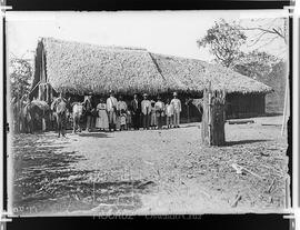 Construção com o telhado em palha, à frente, posando para a foto, homens, mulheres e crianças. À ...