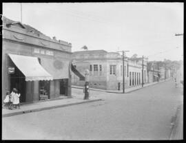 Foto em preto e branco. Uma rua. Na calçada da esquerda, um grupo de meninas, de pé. À direita das meninas, uma loja com um toldo levantado e as portas abertas. Ao lado, outra loja com as portas abertas. Na parede acima das portas, um letreiro: Bar do Norte. À direita, a entrada para uma outra rua. Do outro lado dessa rua, na calçada, várias casas e postes.