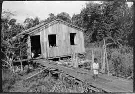 Foto em preto e branco. Área externa com vegetação alta. À direita, sobre uma plataforma de madeira, um menino de pé. Cabelos curtos, camisa de mangas compridas, bermuda e descalço. Atrás da plataforma, à esquerda, uma casa de madeira, com uma porta à esquerda, e duas janelas, à direita. Na porta, duas crianças de pé e um cachorro. Dentro da casa, na janela ao lado da porta, duas crianças de pé. À direita, árvores e vegetação alta. Atrás da casa, árvores.