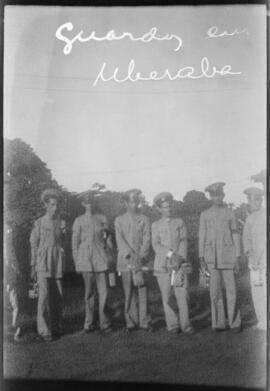 Foto em preto e branco. Numa área externa, um grupo de homens de pé. Todos usam farda: chapéu, paletó com uma braçadeira no braço esquerdo, calça e sapatos