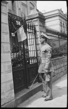 Foto em preto e branco. Uma rua. À esquerda, o portão de uma casa. Fixada ao portão, uma bandeira. À direita, de frente para o portão, um homem de pé, de perfil para a esquerda. O homem usa uniforme: chapéu, paletó com uma braçadeira no lado esquerdo, calça e sapatos. Na mão direita, uma vara. Na mão esquerda, um caderno fechado e um aparelho de aferição: caixa metálica, com uma alça na parte superior.