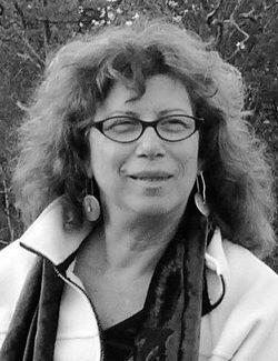 Retrato em preto e branco de Lícia. Ela aparece do busto para cima, com o rosto levemente voltado para a direita. Tem pele clara, cabelos longos, volumosos e ondulados, na altura dos ombros. Usa óculos de armação escura, brincos grandes com pingentes redondos, um casaco claro sobre blusa escura e uma echarpe escura por cima.